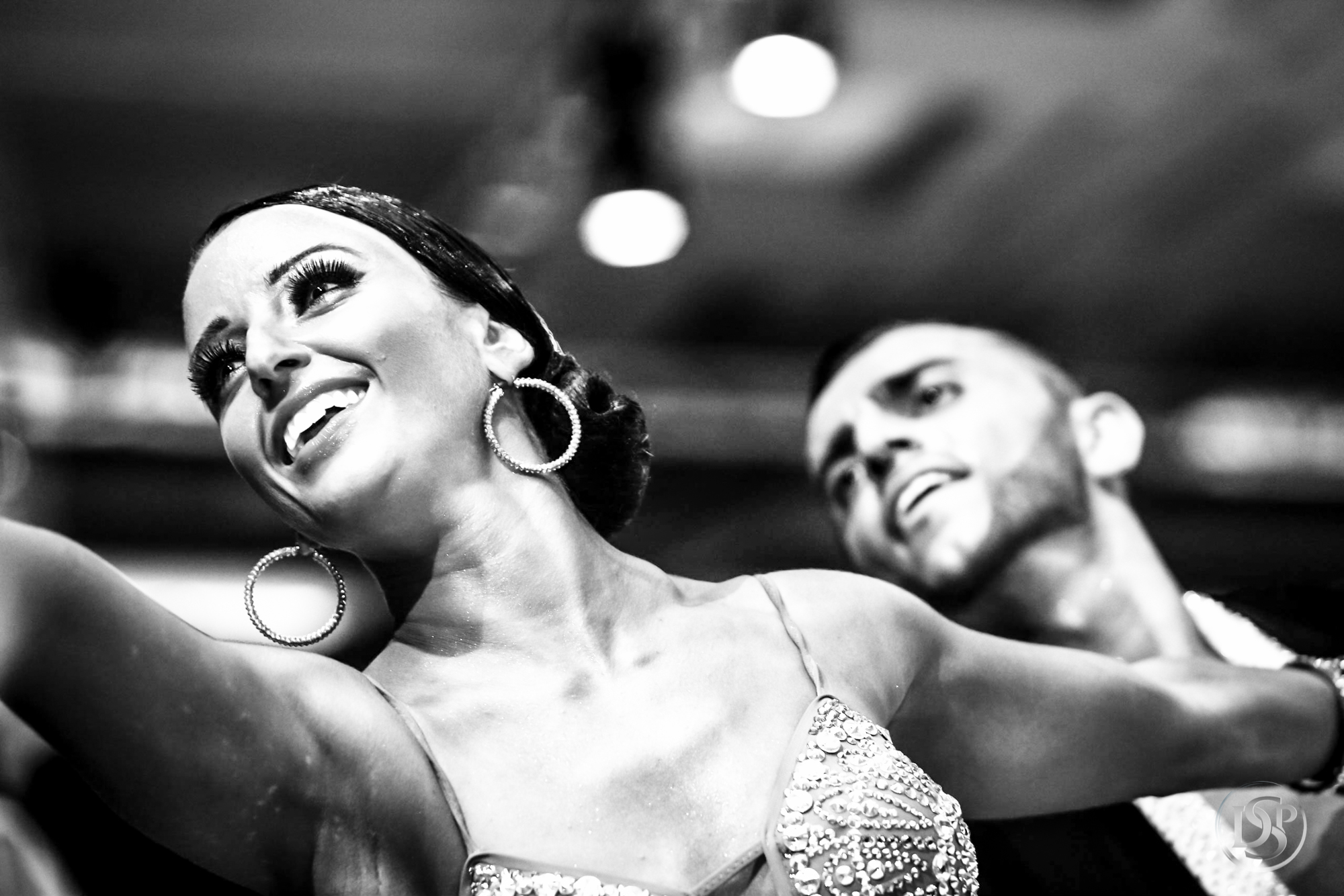 A black-and-white photograph of two dancers in an open position, heads turned to the line of dance under the ballroom lights